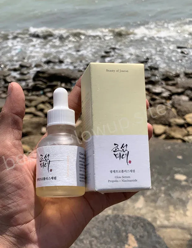Hand holding a skincare serum bottle and its packaging with a beach background