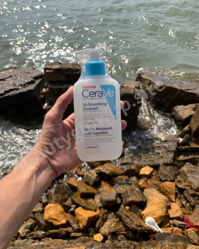 Person holding a bottle of CeraVe SA Smoothing Cleanser by a rocky shoreline.