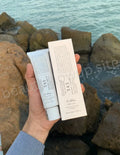 Hand holding skincare products with a scenic background of rocks and water.