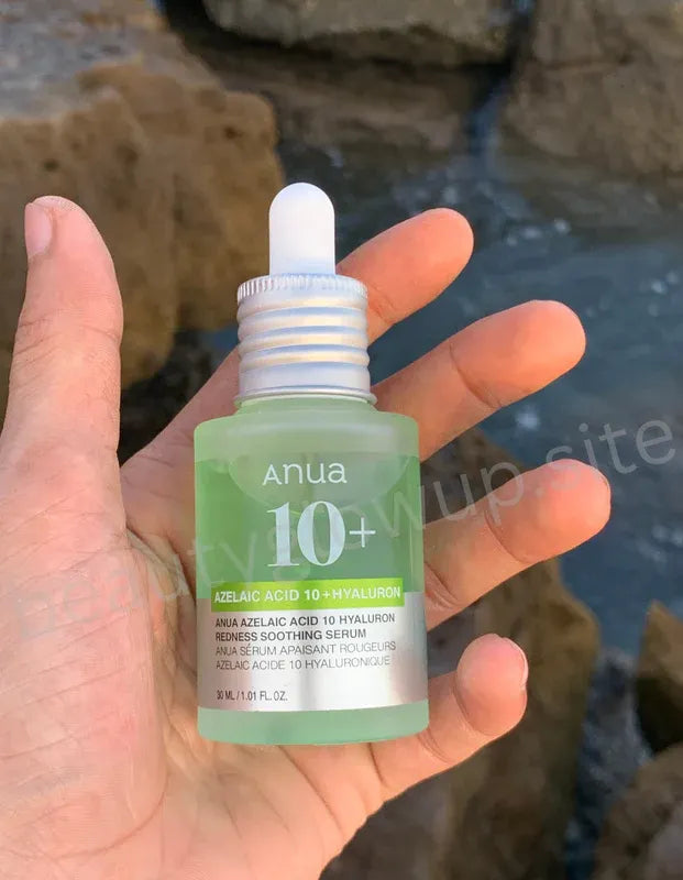 Hand holding a green skincare serum bottle labeled 'anua 10+' with a blurred natural background.