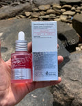 ANUA Niacinamide 10 percent TXA 4 percent Dark Spot Correcting Serum 30ml Hand holding a red serum bottle with dropper and its packaging against a rocky background